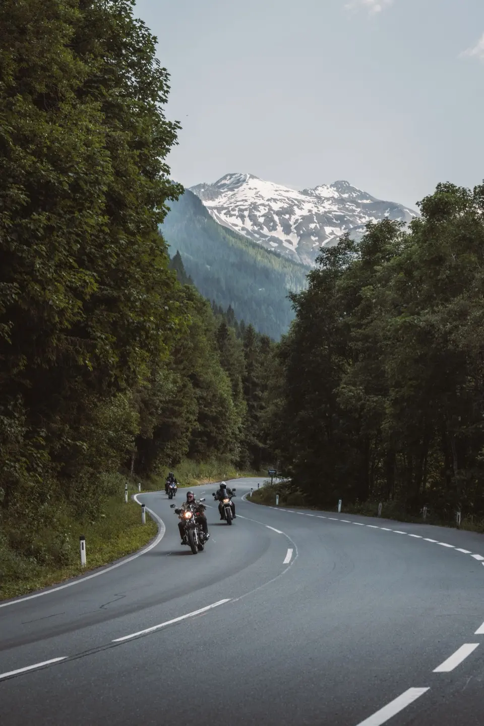 Three motorcycles ride along a country road through the forest, a snow-capped mountain behind them